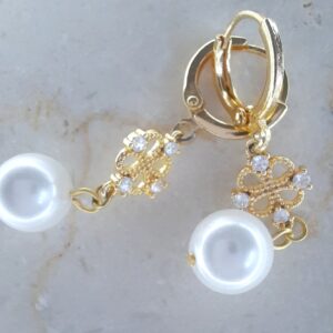 Add a touch of refined romance to your look with the Fleur de Perle Drop Earrings. These elegant earrings feature a gleaming gold-tone hoop, adorned with a delicate filigree clover motif set with sparkling white crystals. Suspended below, a luminous artificial pearl adds a classic finish, creating a harmonious blend of vintage charm and modern sophistication.